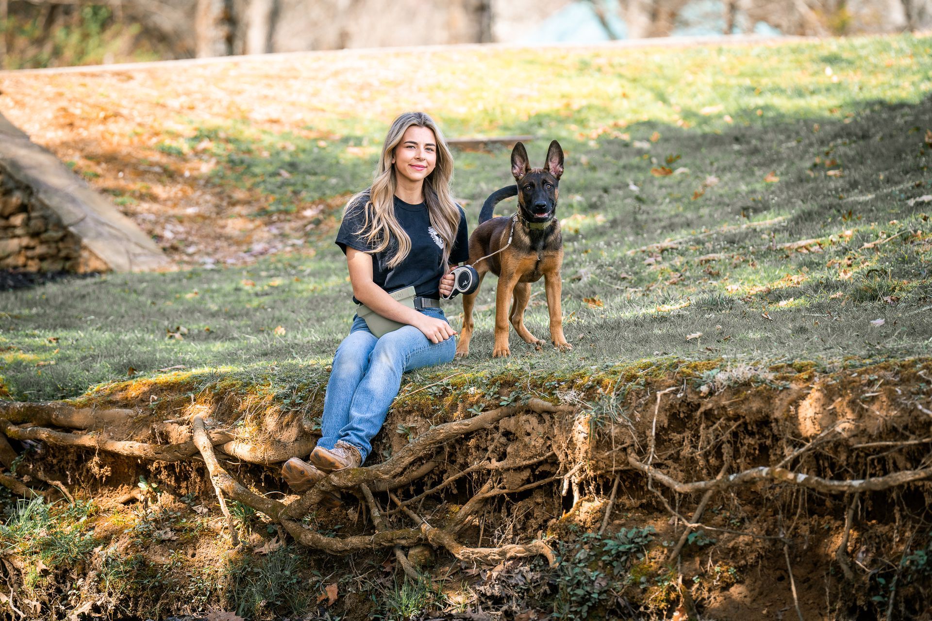 A woman sits on an earthen bank with a Belgian Malinois dog standing beside her outdoors on a sunny, grassy hillside.