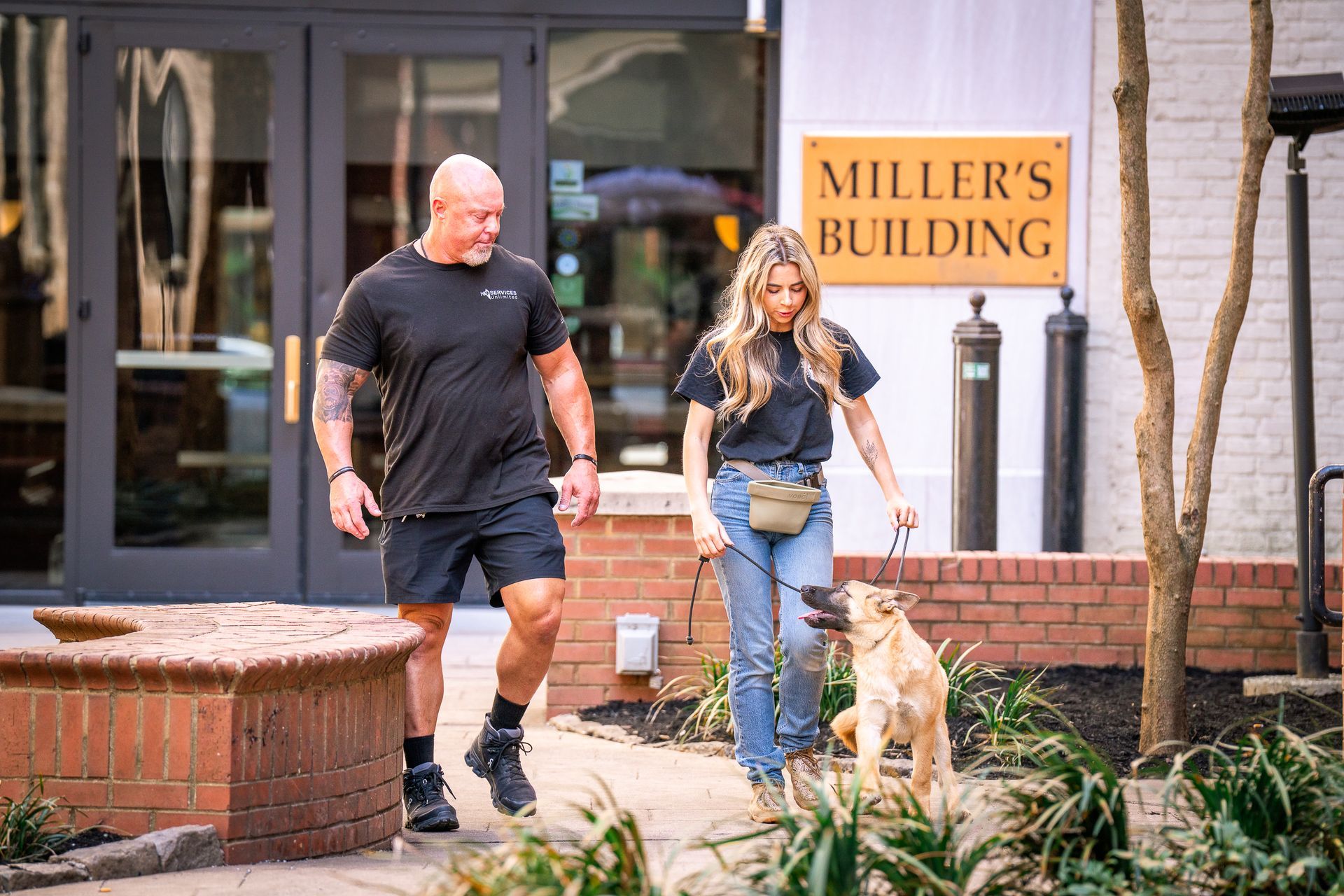 A muscular person and a woman with a leashed dog walking outside the Miller's Building on a brick-lined path.