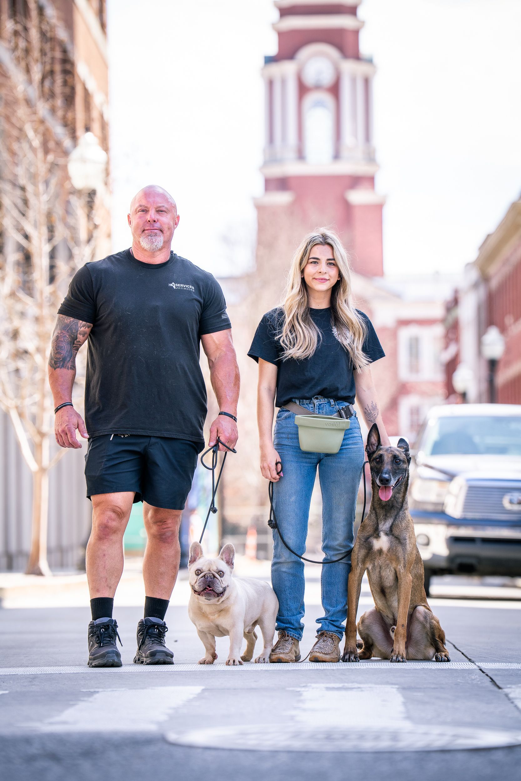 A person and a partner standing on a crosswalk with a French bulldog and a Belgian Malinois, with a clock tower behind.