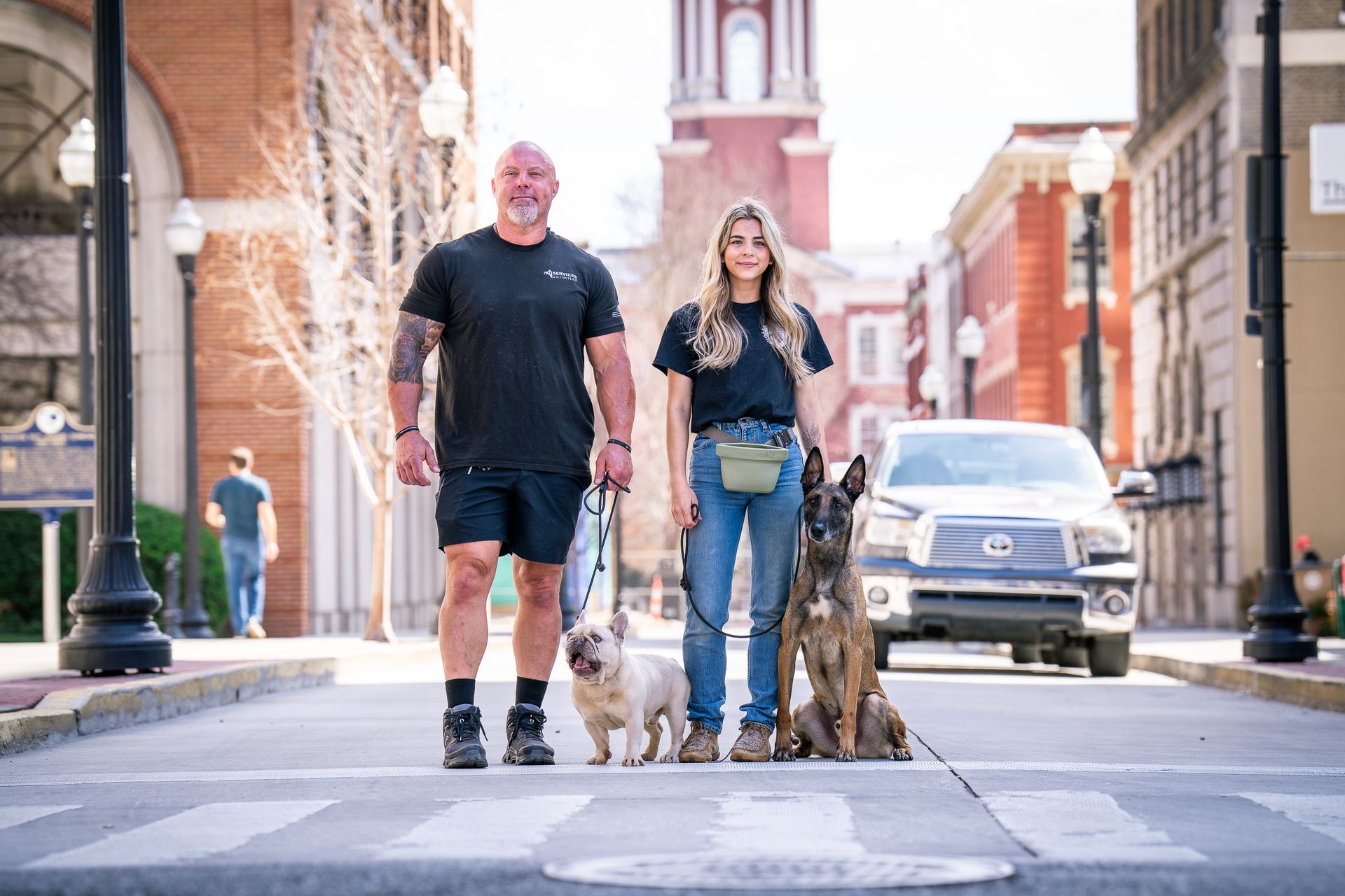 A person and their companion stand on a city street with a French Bulldog and a Belgian Malinois on leashes.