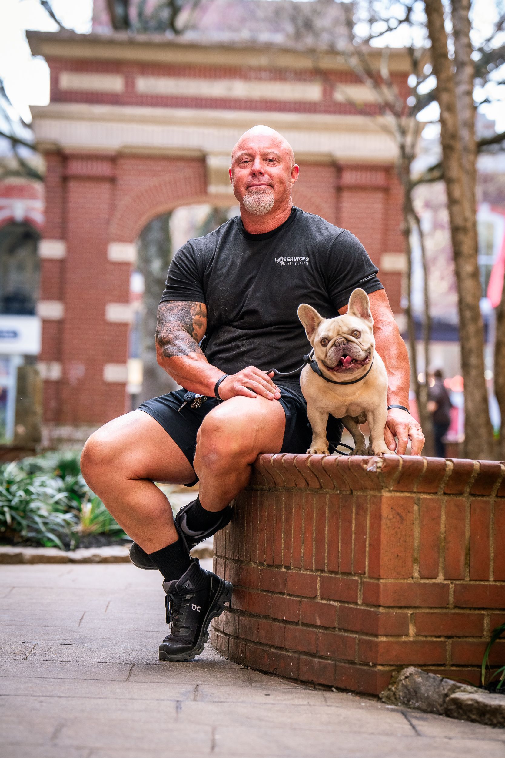 A muscular person sits on a brick wall beside a fawn-colored French Bulldog in an outdoor plaza with a brick archway.