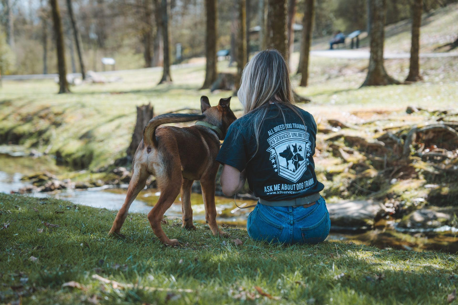 A person with long wavy hair sits on a grassy bank by a stream with a medium-sized brown dog standing next to them.