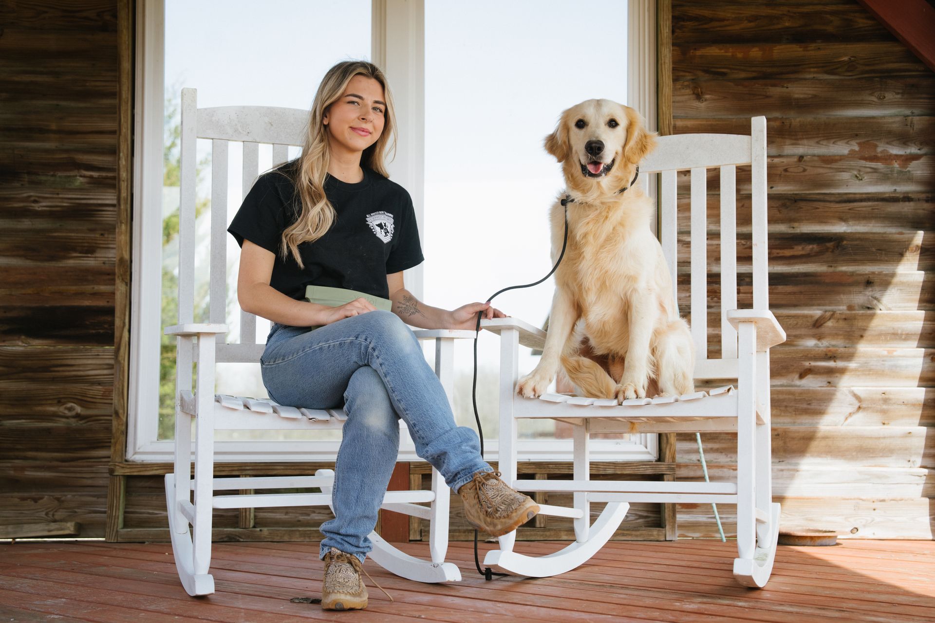 A person sitting on a white rocking chair next to a golden retriever sitting on another, on a porch with wooden walls.