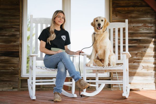 A person sitting on a white rocking chair next to a golden retriever sitting on another, on a porch with wooden walls.