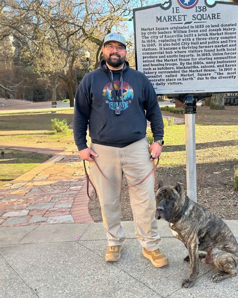 A person wearing a hoodie and hat stands on a path with a brindle dog, next to a historic Market Square sign.