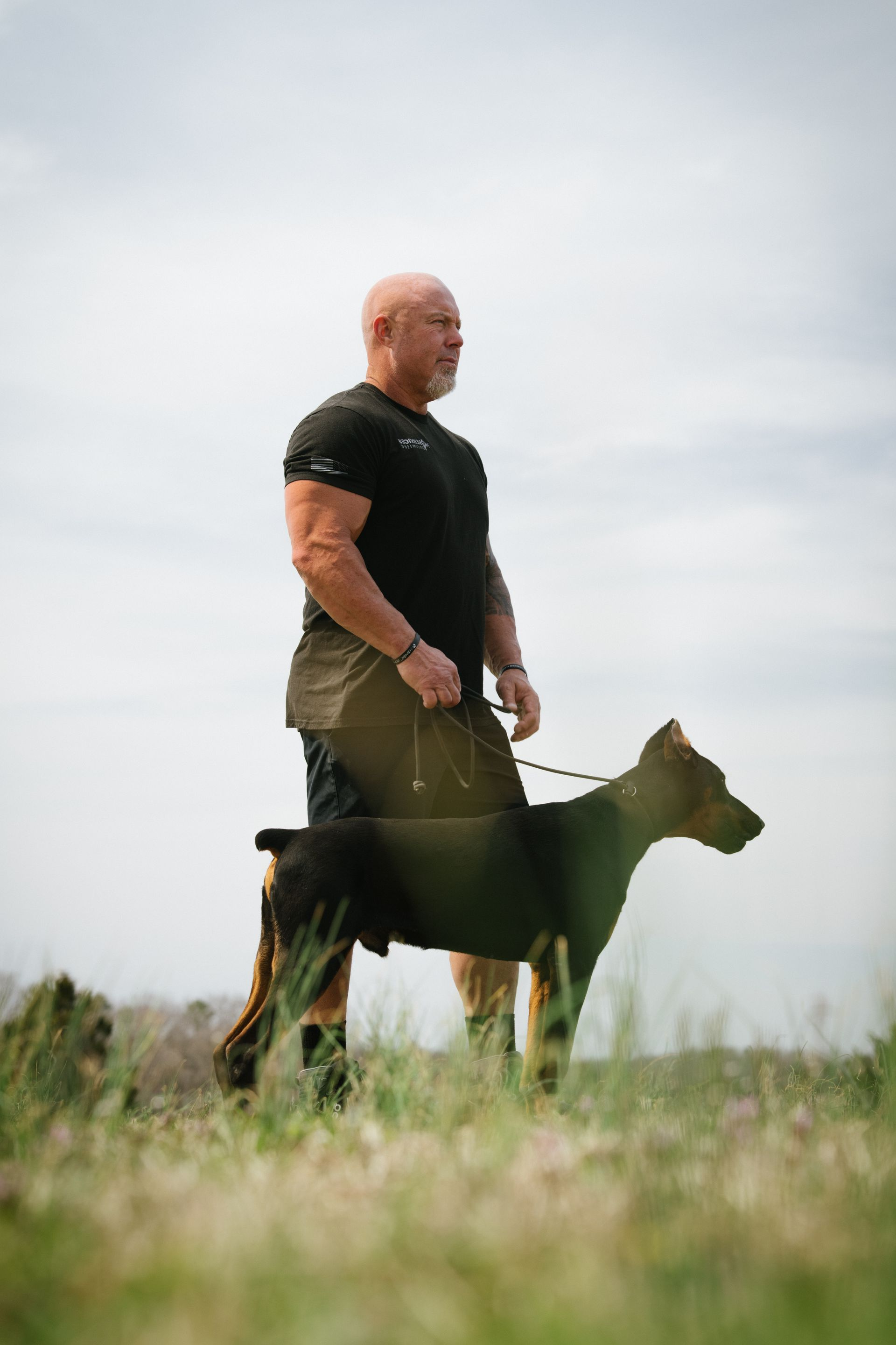 A bald, muscular person stands in a grassy field, holding a leash attached to a standing Doberman.