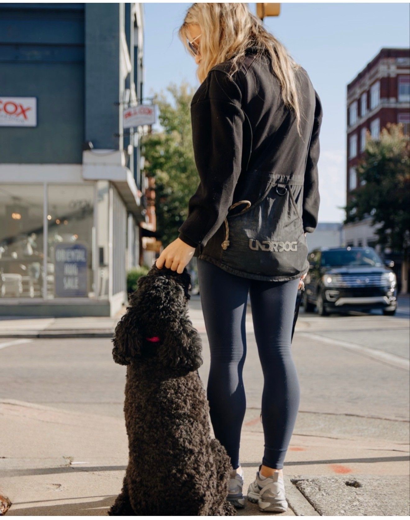 A person in a black jacket and leggings stands on a city sidewalk holding a black dog’s leash.
