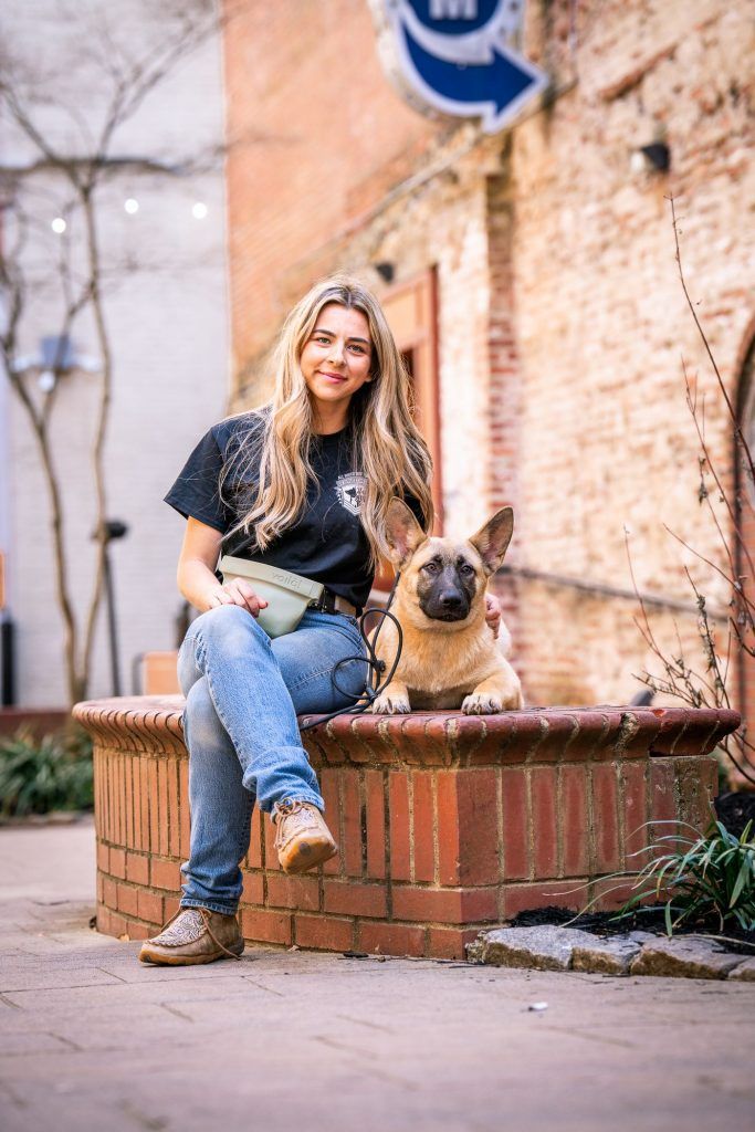A person with long wavy hair sits on a brick ledge outdoors next to a tan shepherd-type dog, both looking at the camera.