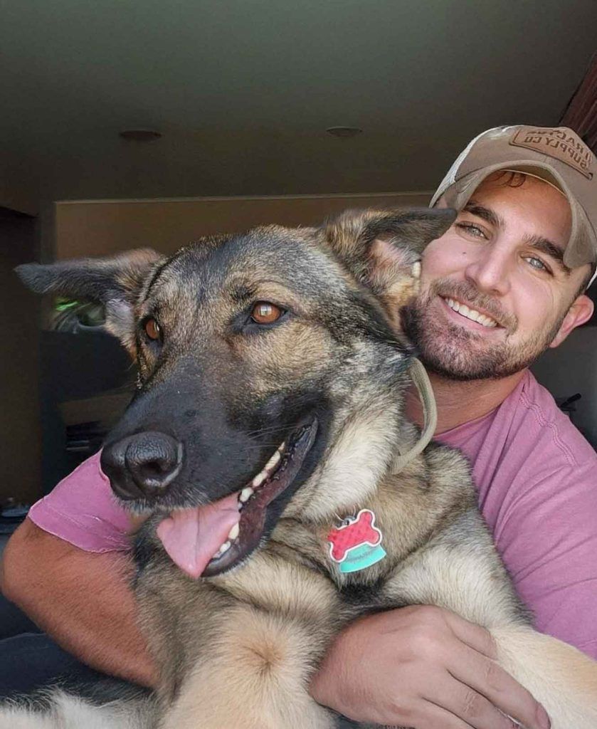 A person in a baseball cap smiles while holding a German Shepherd with an open-mouthed expression.