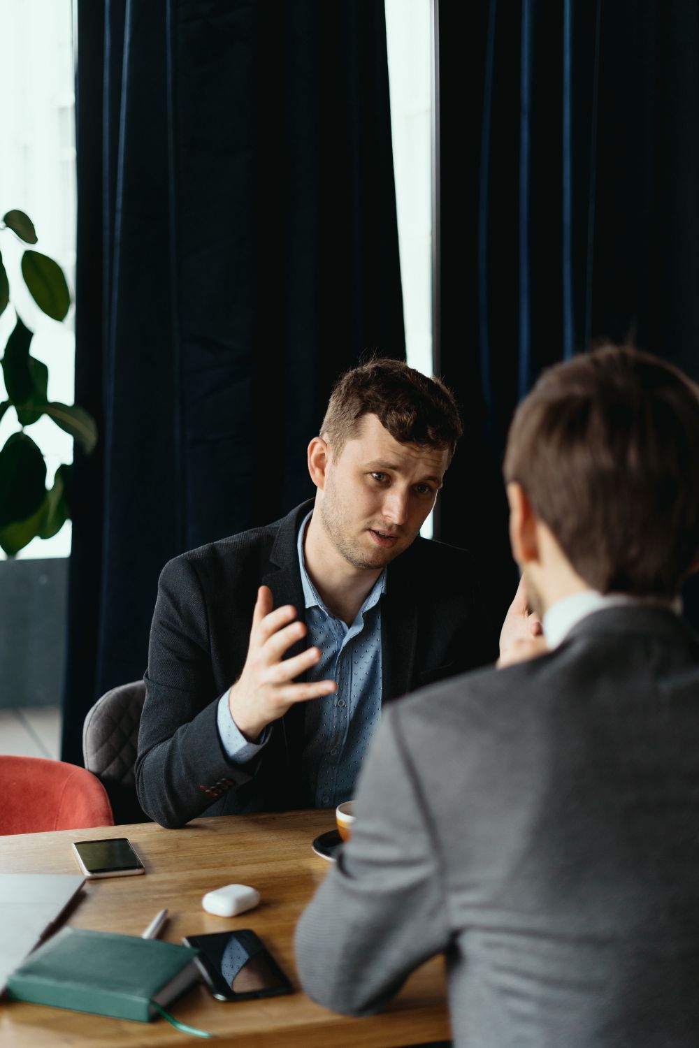 Two people in a business meeting across a wooden table, one gesturing while speaking near a window.