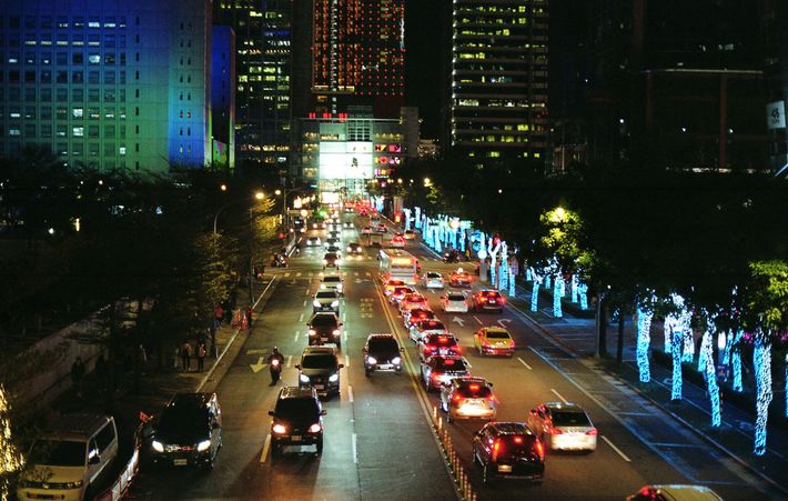 Nighttime city street with cars in traffic and blue-lit trees along the road