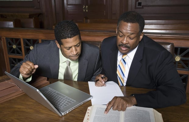 Two men are sitting at a table with a laptop and a book.