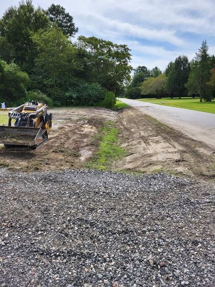 Skid steer removing vegetation beside a road, grading a gravel parking area. Trees and cloudy sky in background.