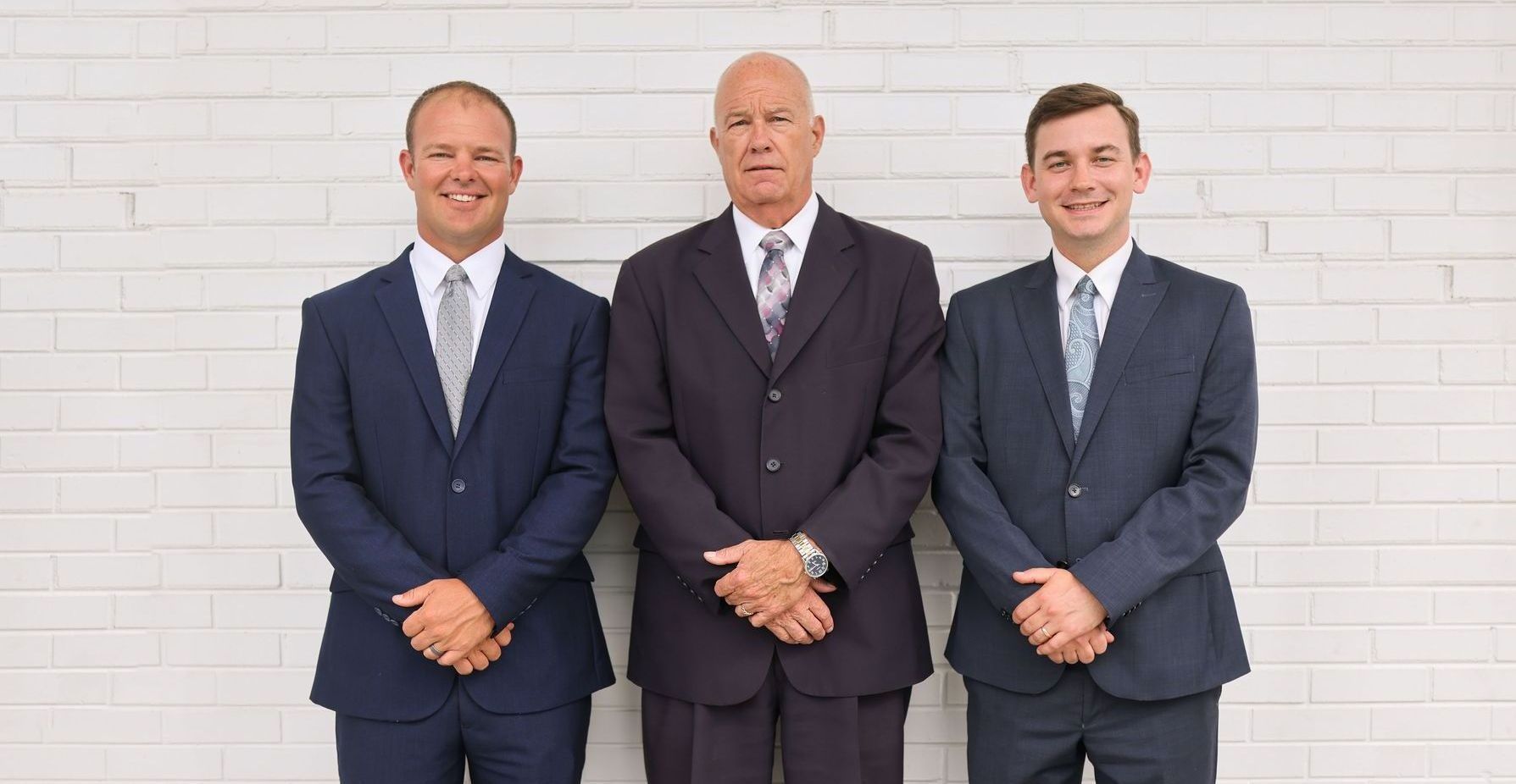 Three men in suits and ties are standing next to each other in front of a white brick wall.