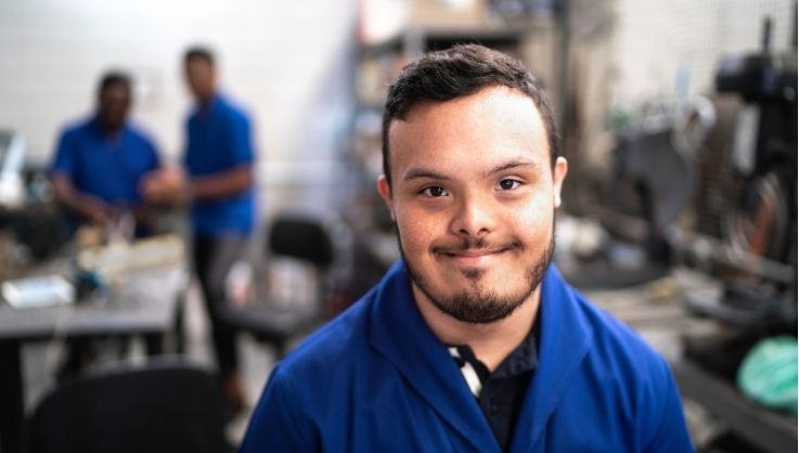 A man with down syndrome is standing in a factory and smiling at the camera.