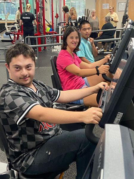 A man is helping another man on a rowing machine in a gym.