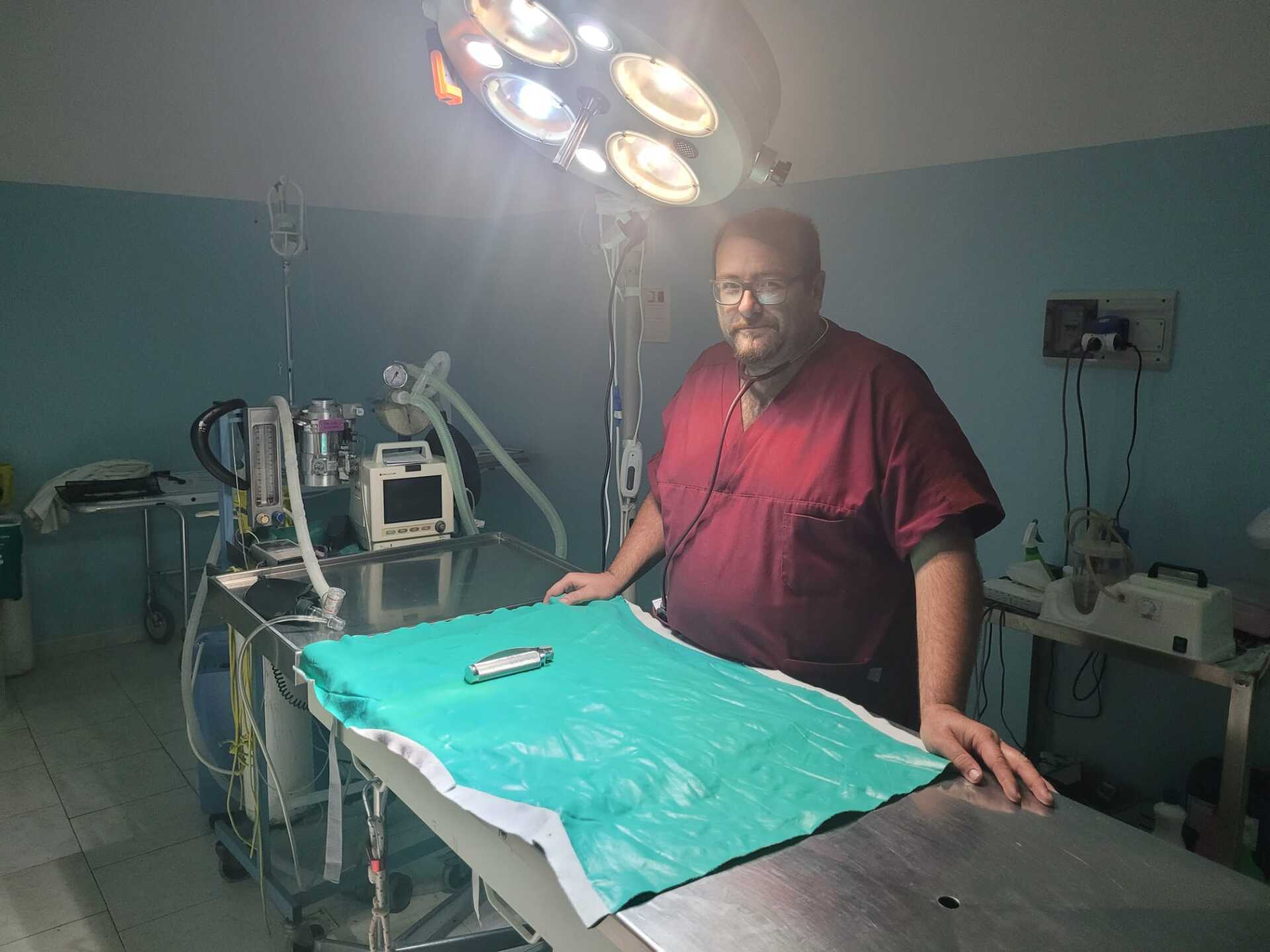 Una persona in camice bordeaux è in piedi in una sala operatoria, di fronte alla telecamera. In primo piano c'è un tavolo operatorio con una copertura verde.