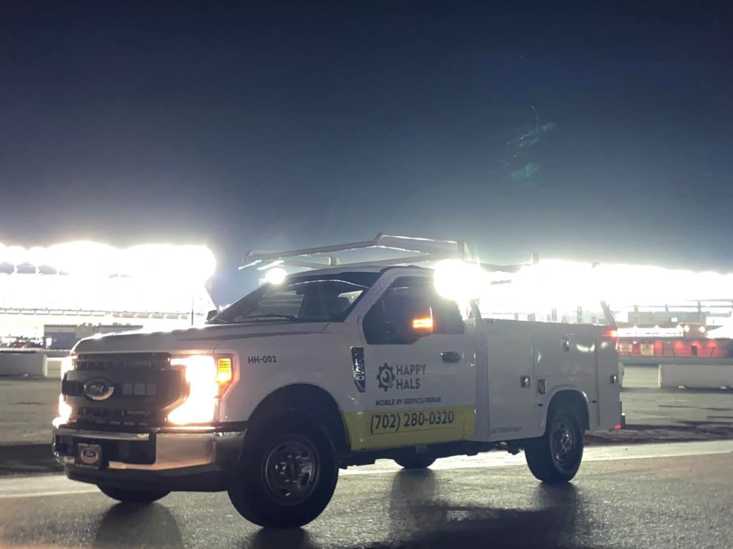 White work truck with lights on, parked in a wet area under a cloudy, dark sky.