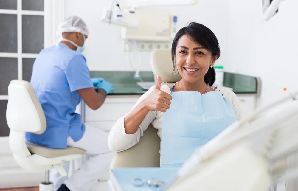 A woman is sitting in a dental chair giving a thumbs up.