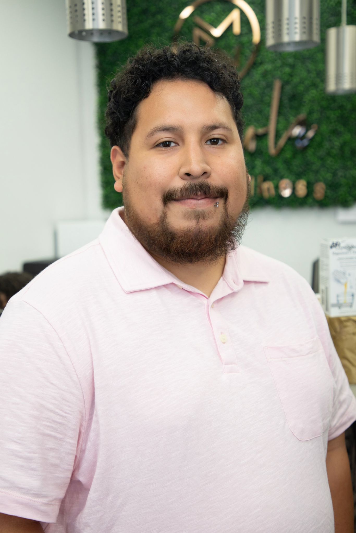 A man with a beard and a pink shirt is standing in front of a green wall.