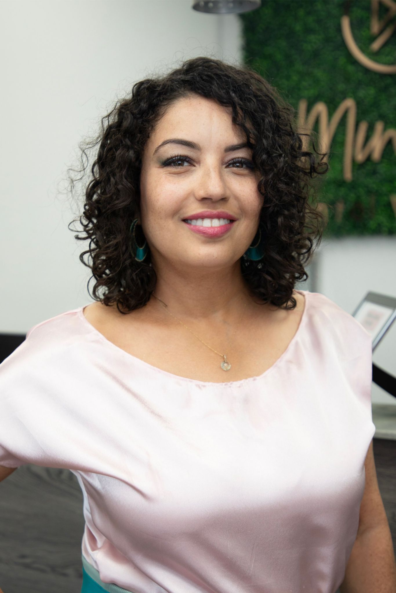A woman with curly hair is wearing a pink shirt and smiling.