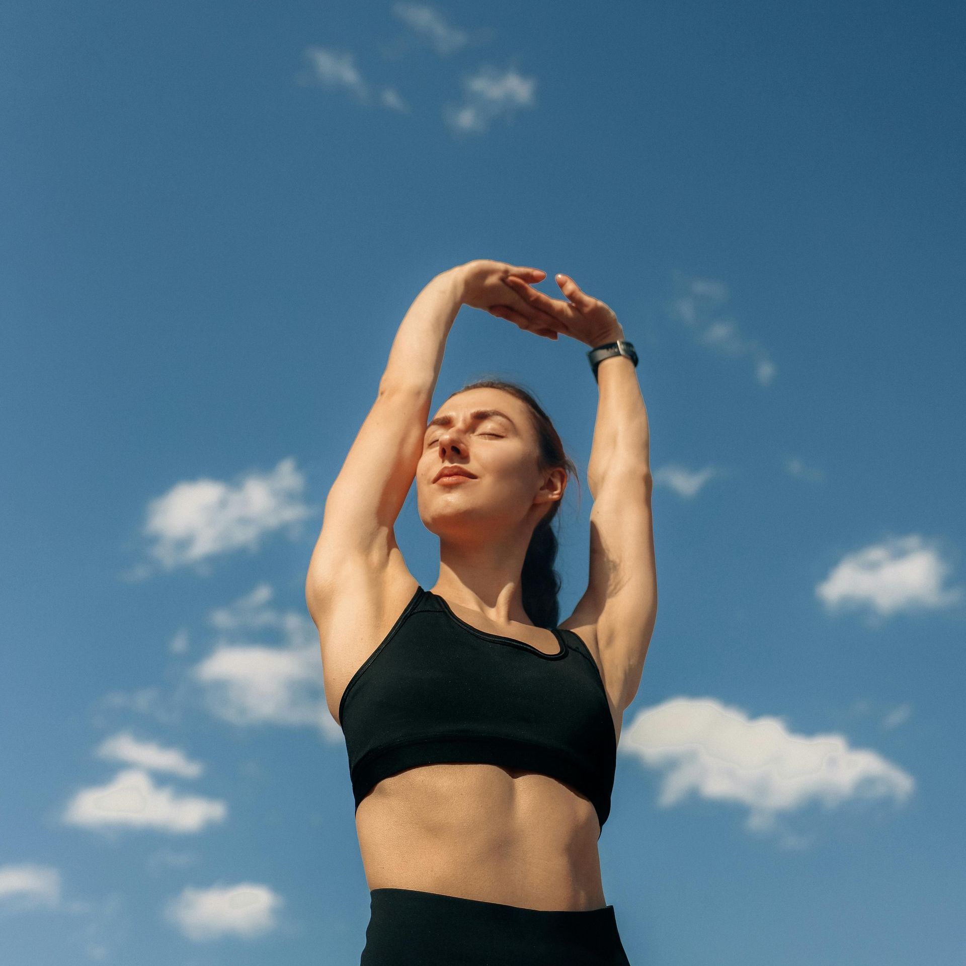 A woman in a black crop top is stretching her arms in front of a blue sky