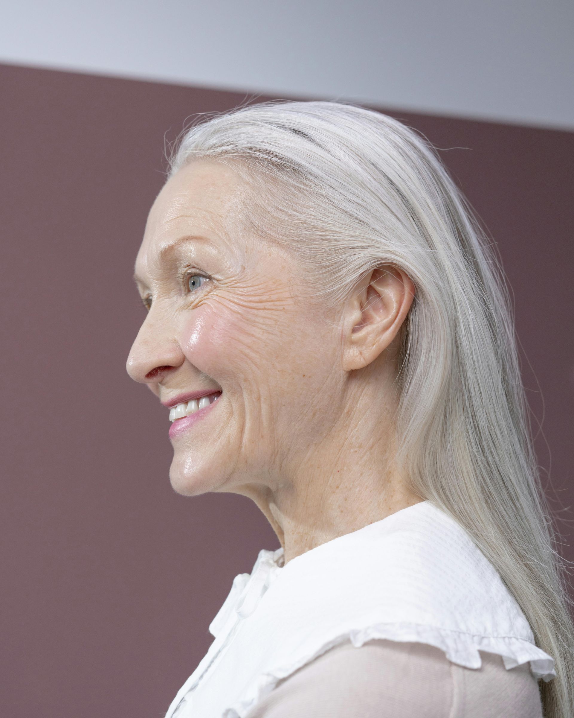 A woman with long gray hair is smiling and wearing a white shirt.