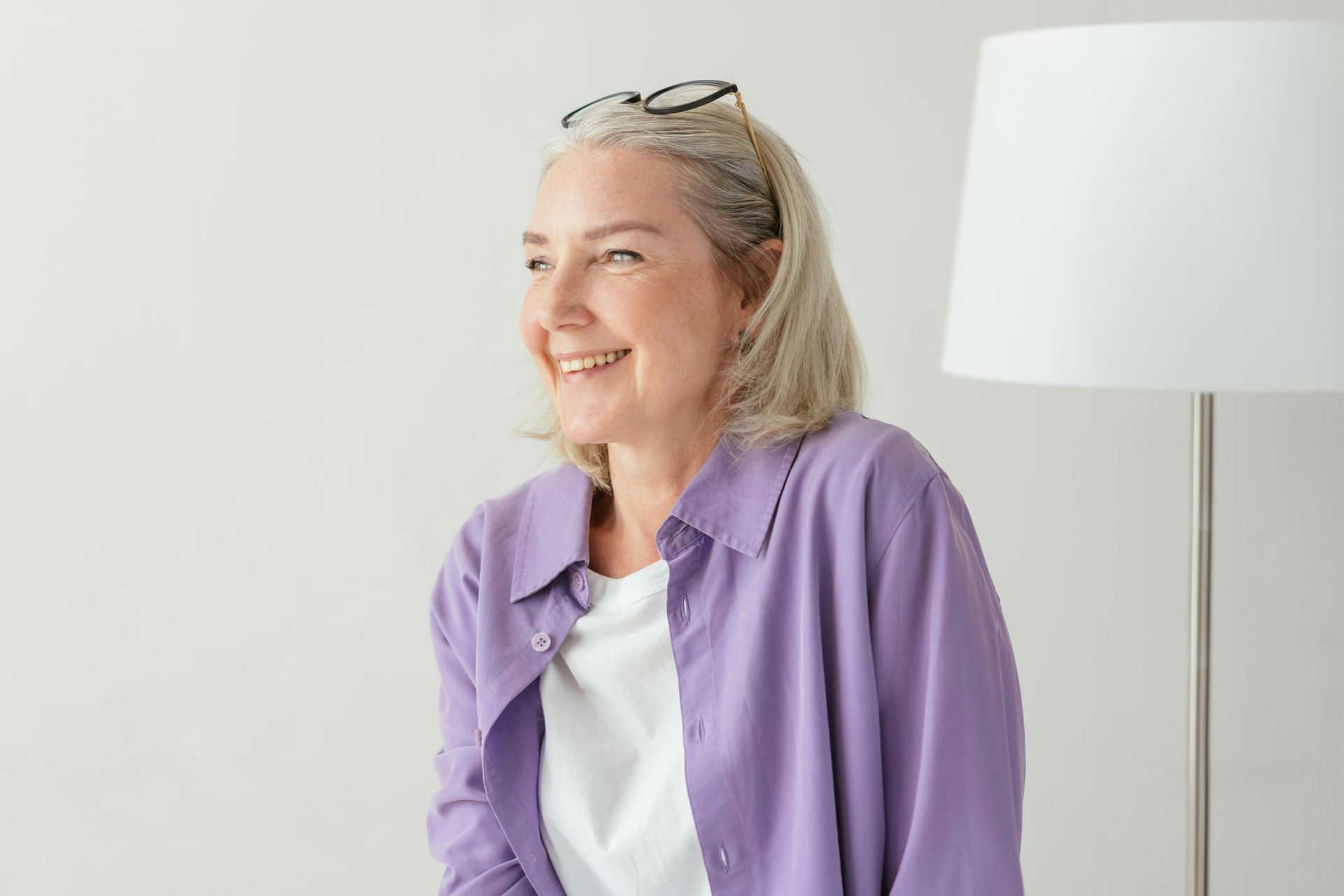 An older woman wearing glasses and a purple shirt is sitting in front of a lamp.