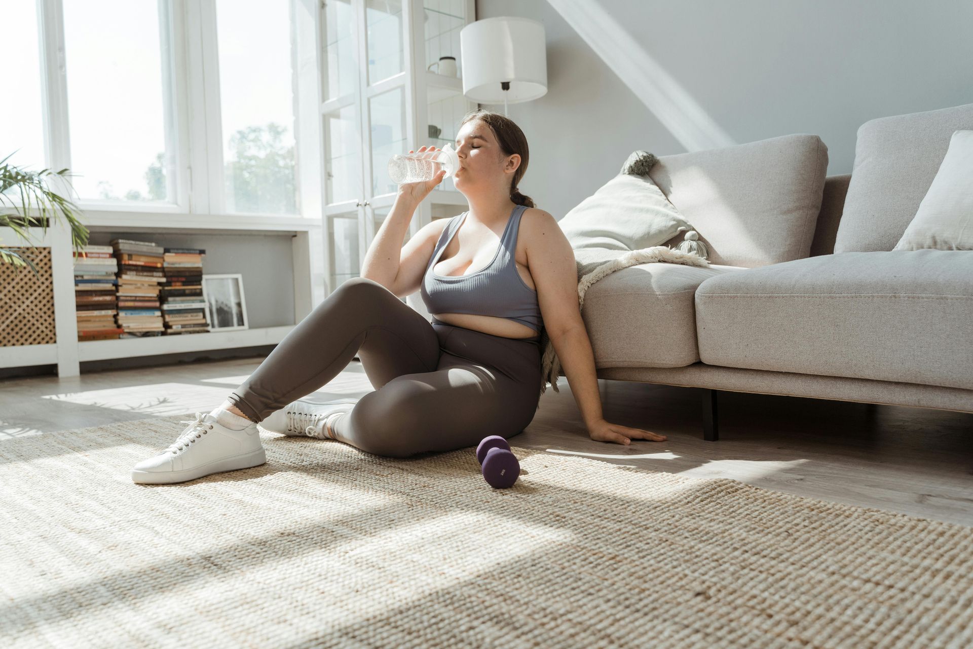 A woman is sitting on the floor drinking water in a living room.