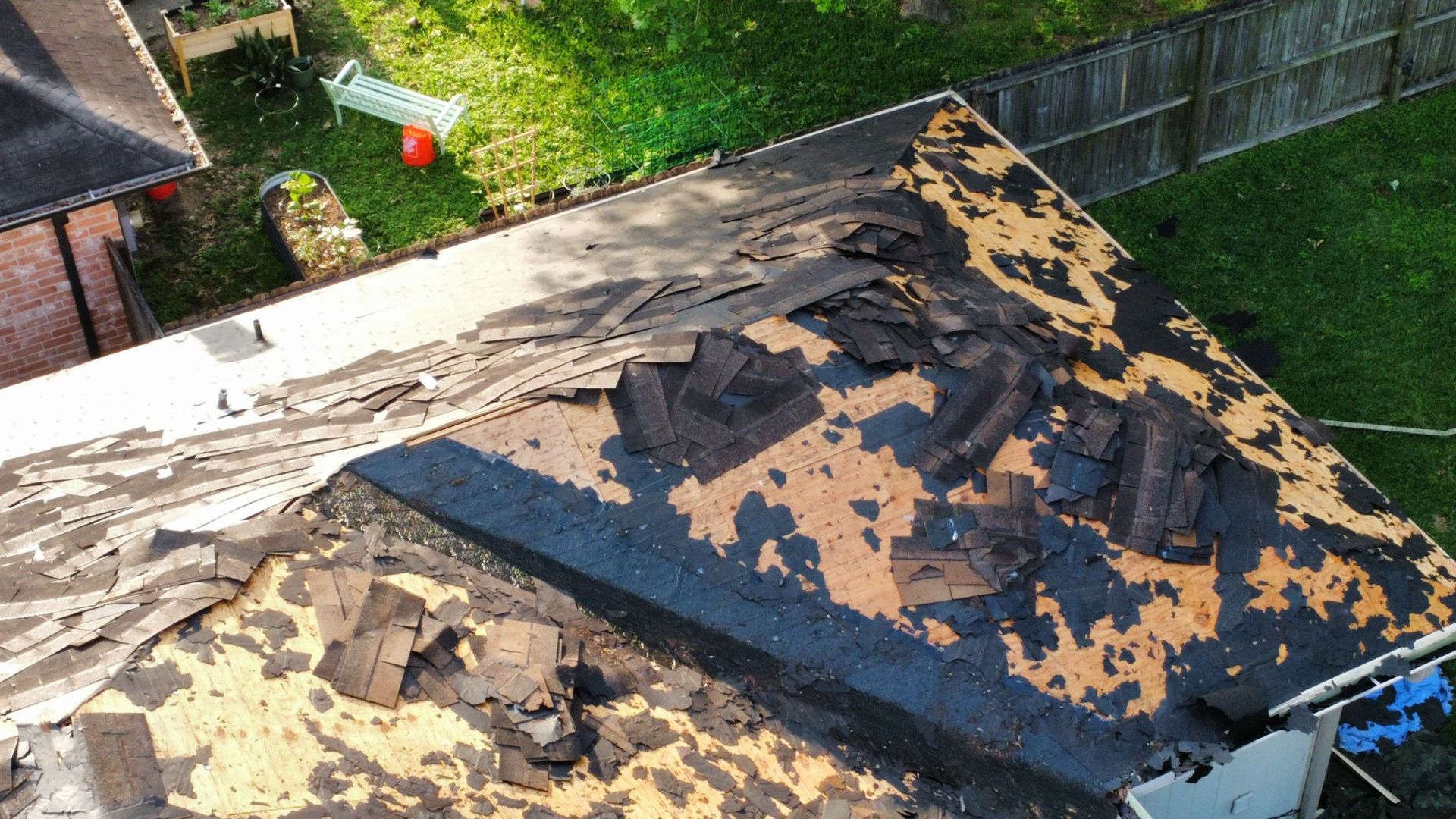 Damaged roof with torn black shingles and exposed underlayment beside a fenced backyard