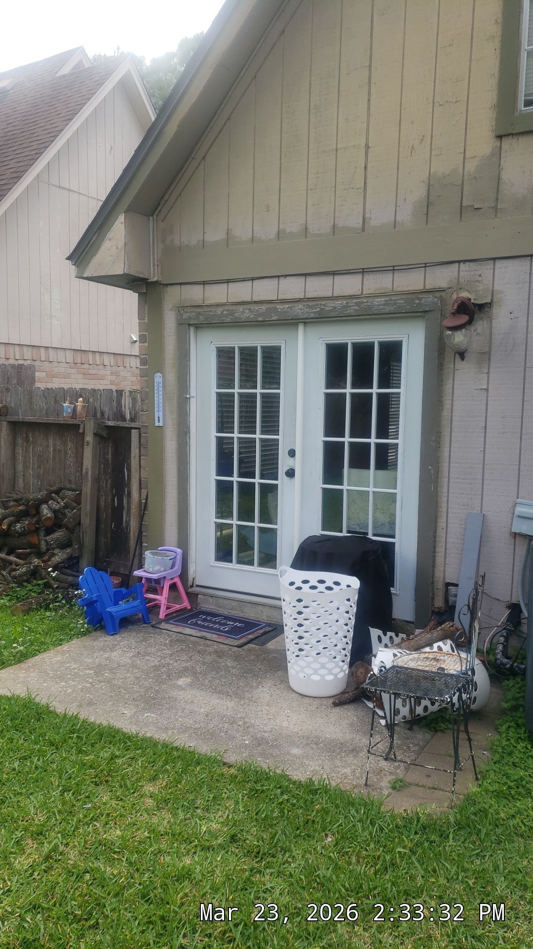 Back patio with white French doors, trash bin, chairs, and a small table beside a beige house.