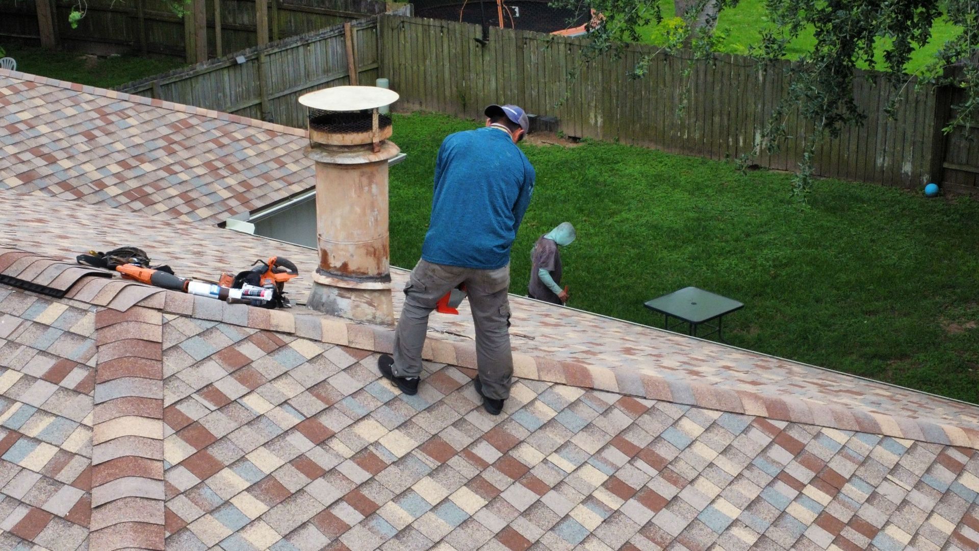 Worker on a sloped tiled roof near a chimney, carrying a tool by a grassy backyard.