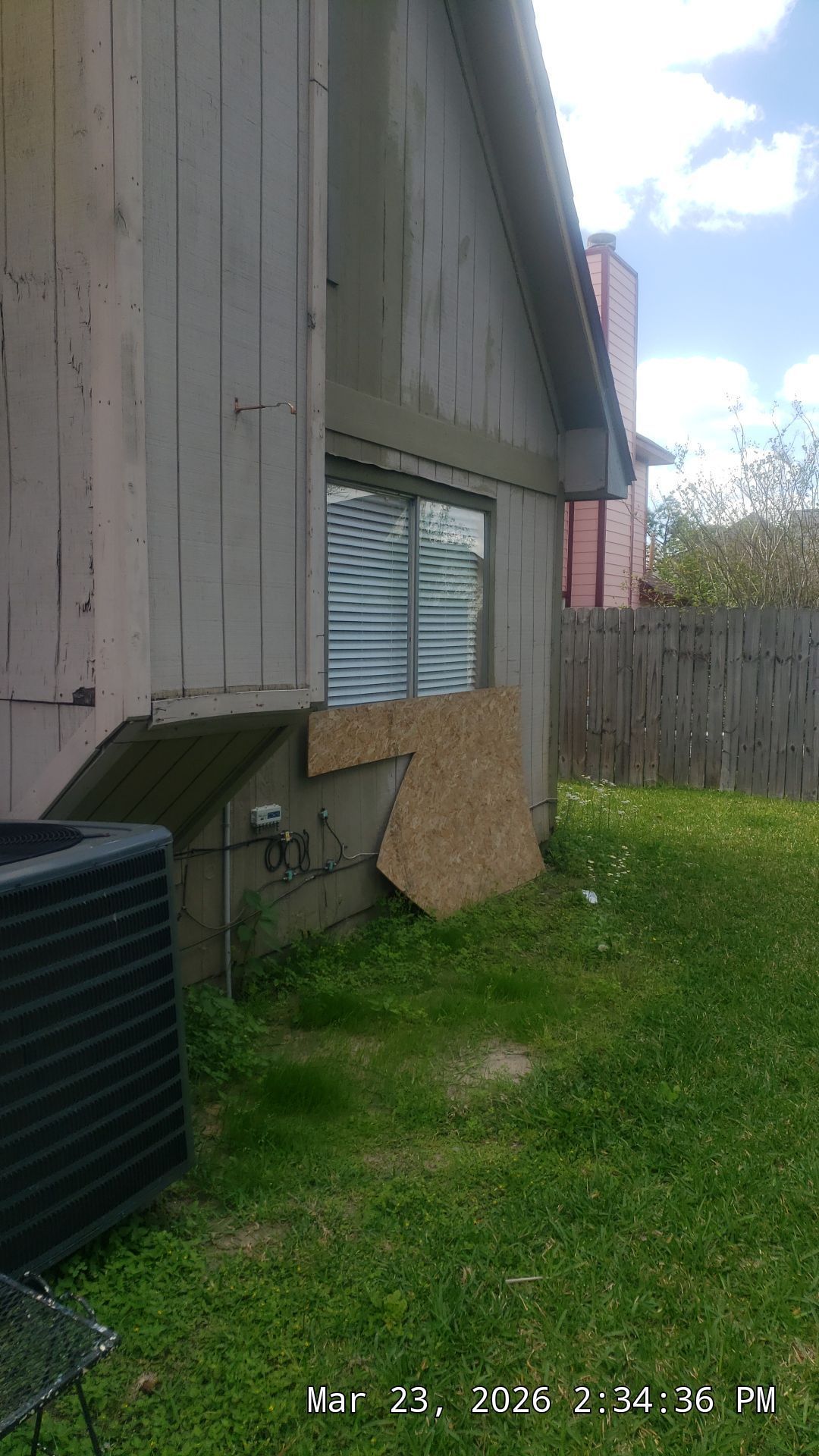 Side of a weathered house with a basement window, air conditioner, and grassy yard beside a wooden fence.