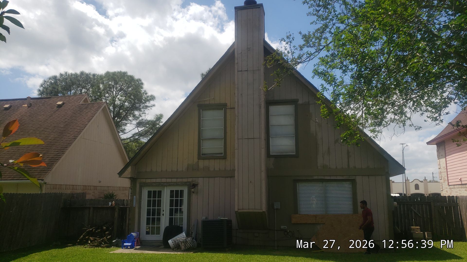 A tan house with a steep roof and central chimney, front porch, and trees in the yard.
