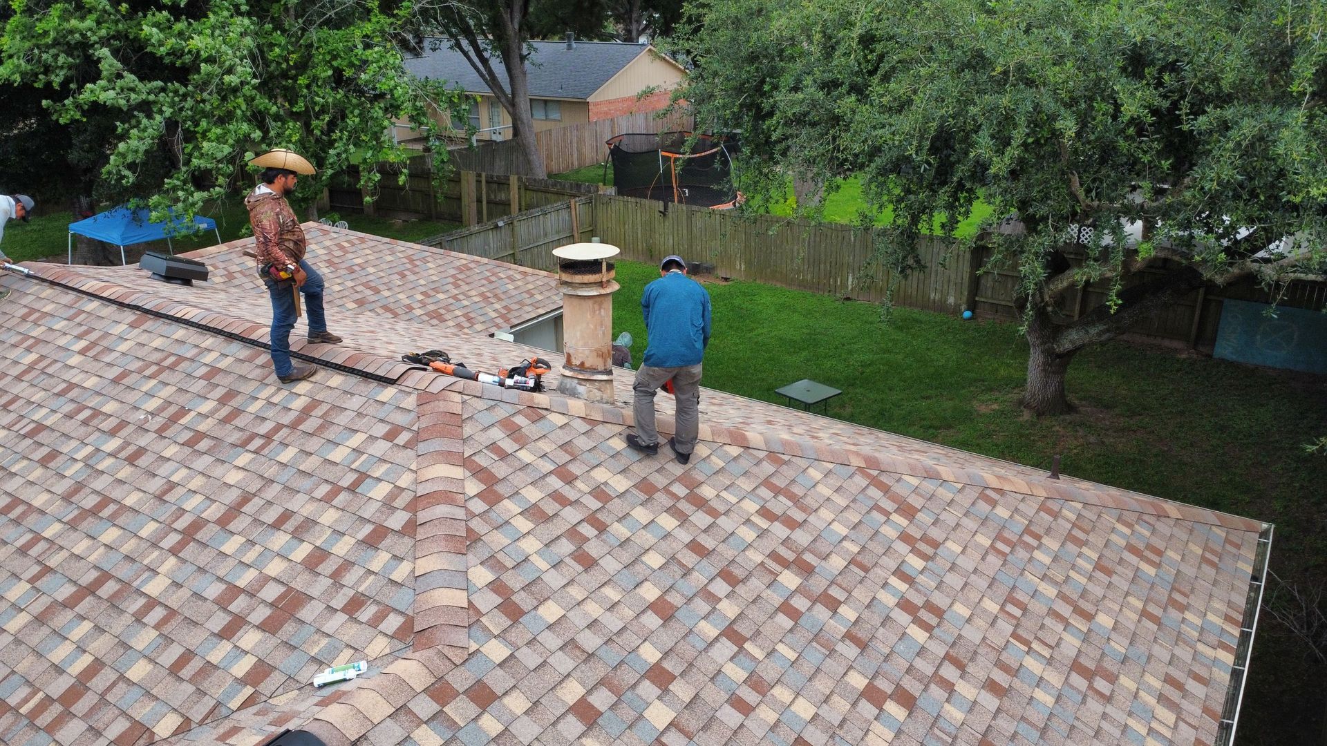 Two workers on a brick roof near trees, one standing by tools and a vent pipe.