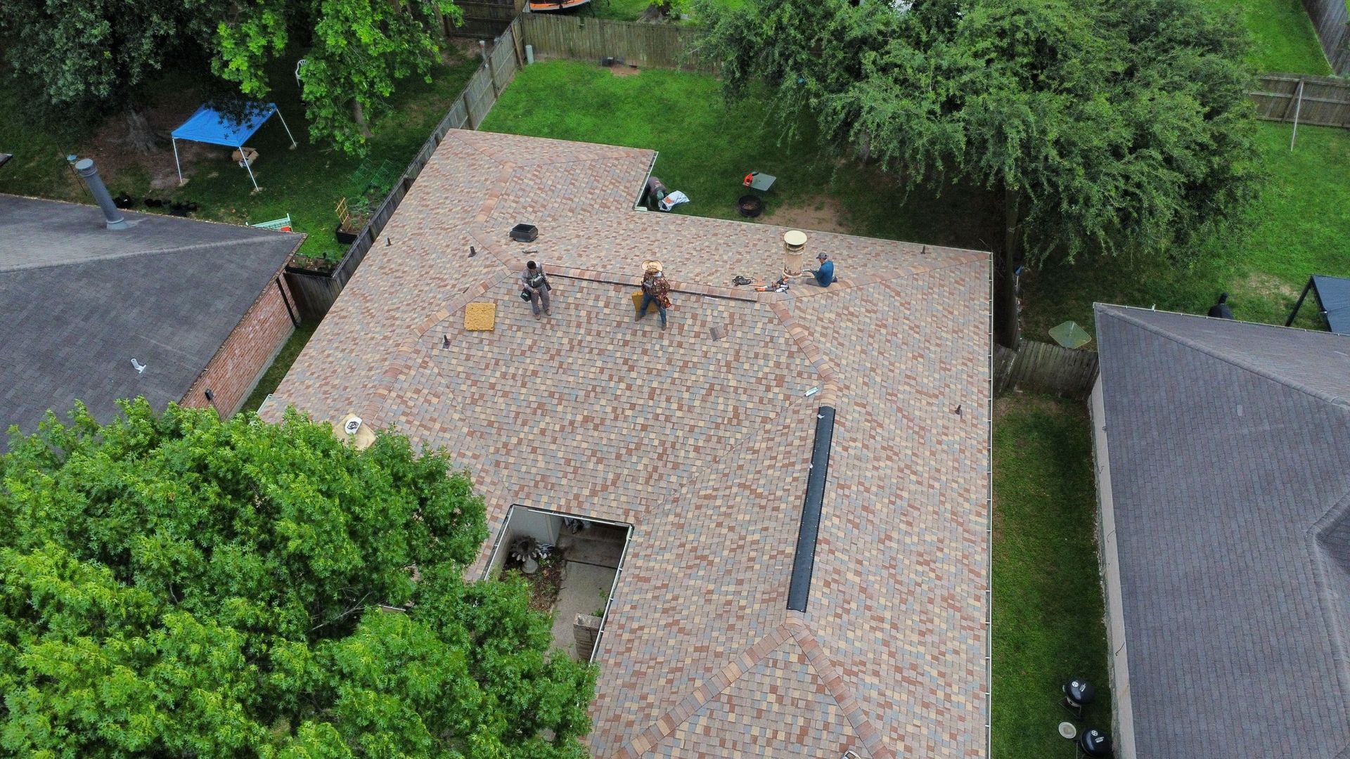 Aerial view of a shingled roof with several workers near a roof vent, surrounded by trees and neighboring houses