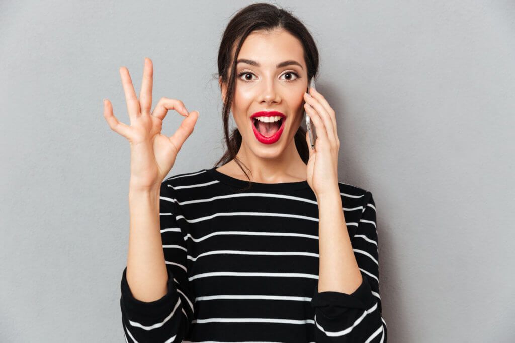 A woman is making an ok sign while talking on a cell phone.