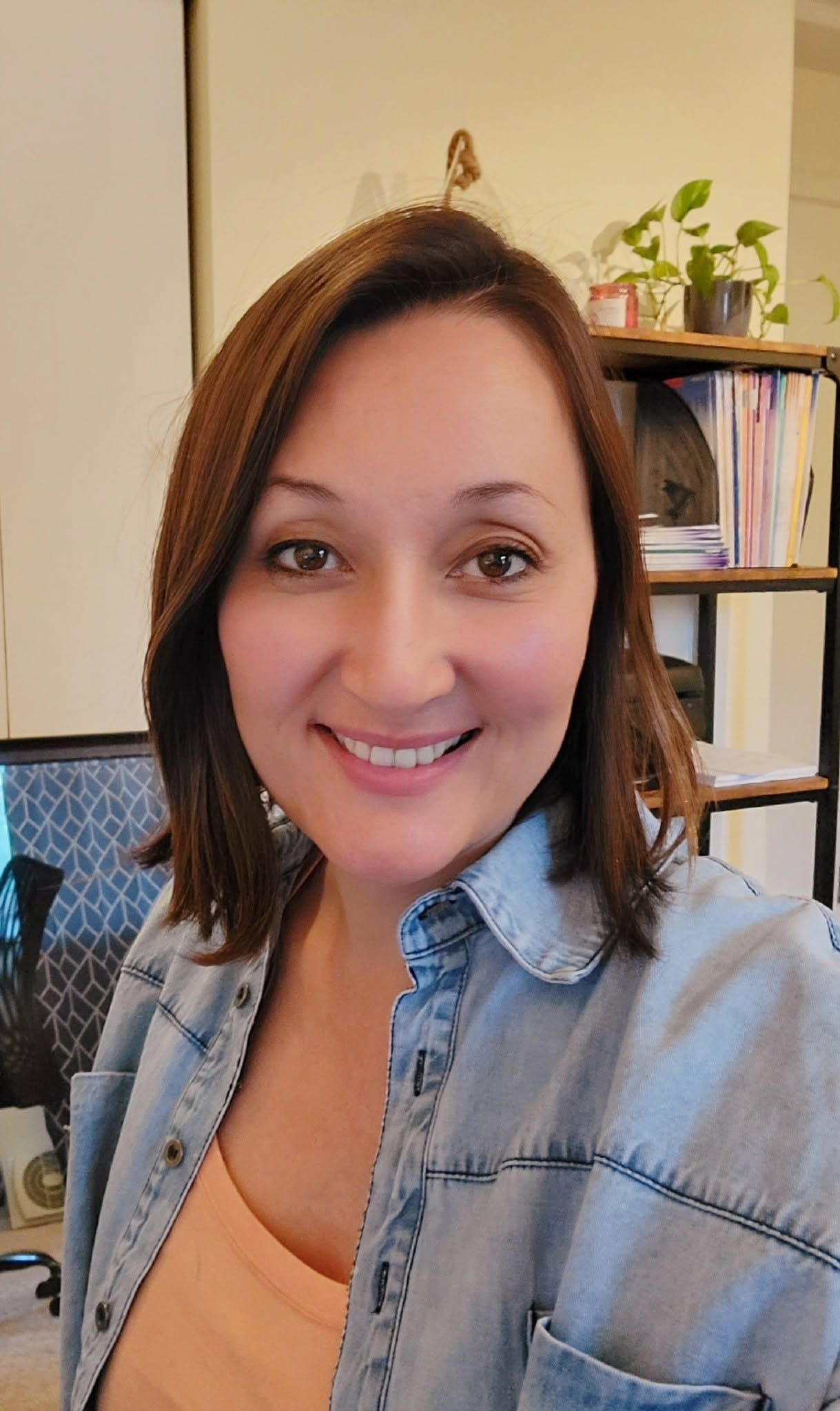 Smiling woman in a denim shirt taking a selfie indoors beside a bookshelf and potted plant.