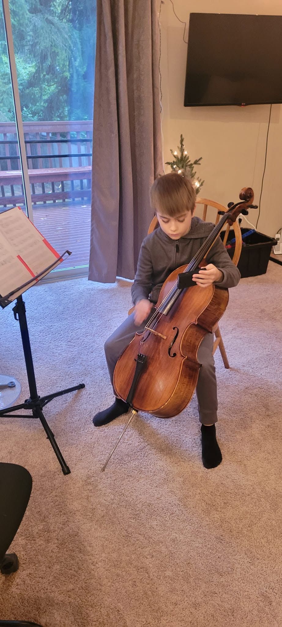 Child playing cello in a living room with music stand and TV in the background