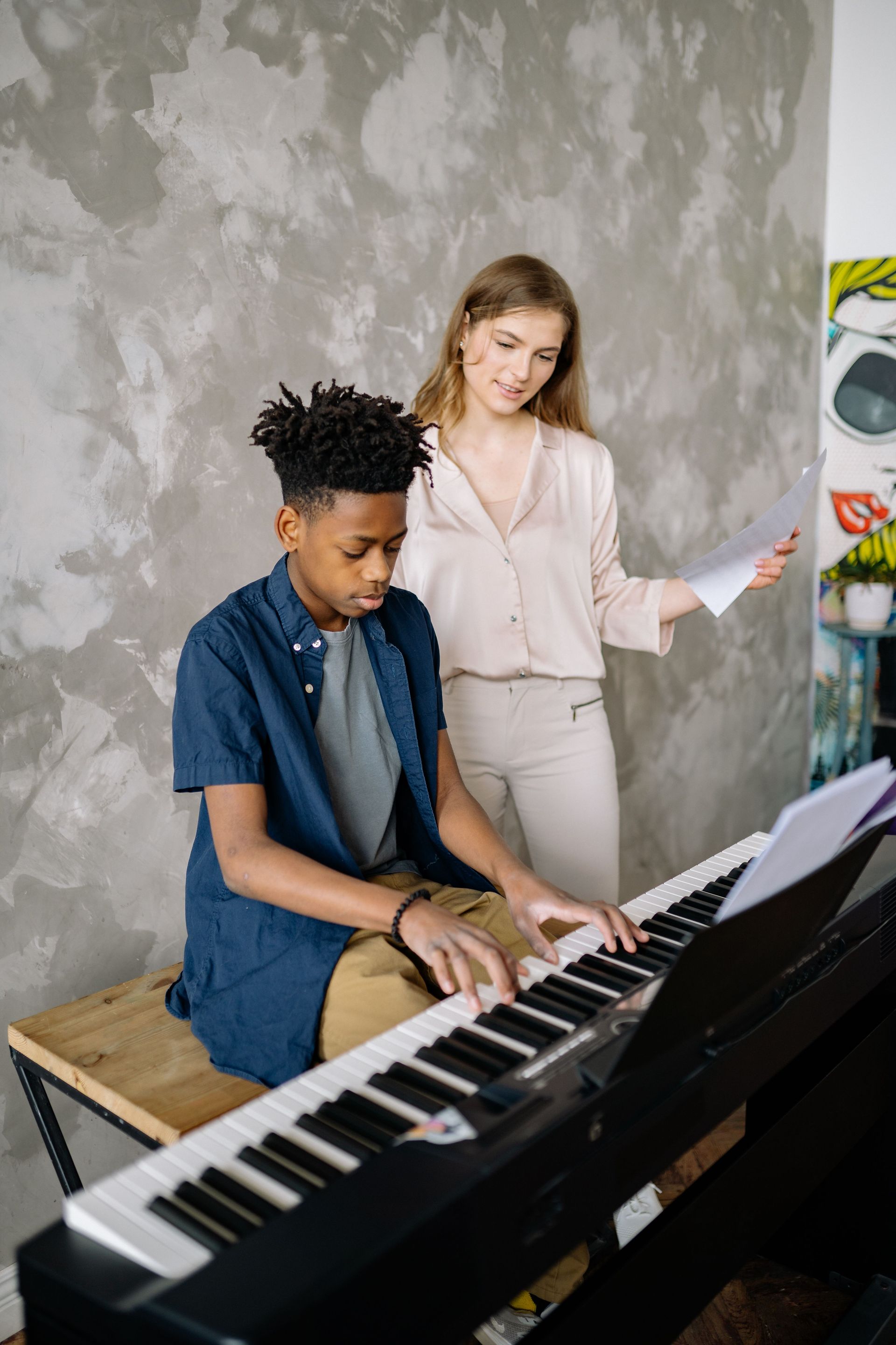Person playing keyboard while another stands nearby in a music room with a gray textured wall.