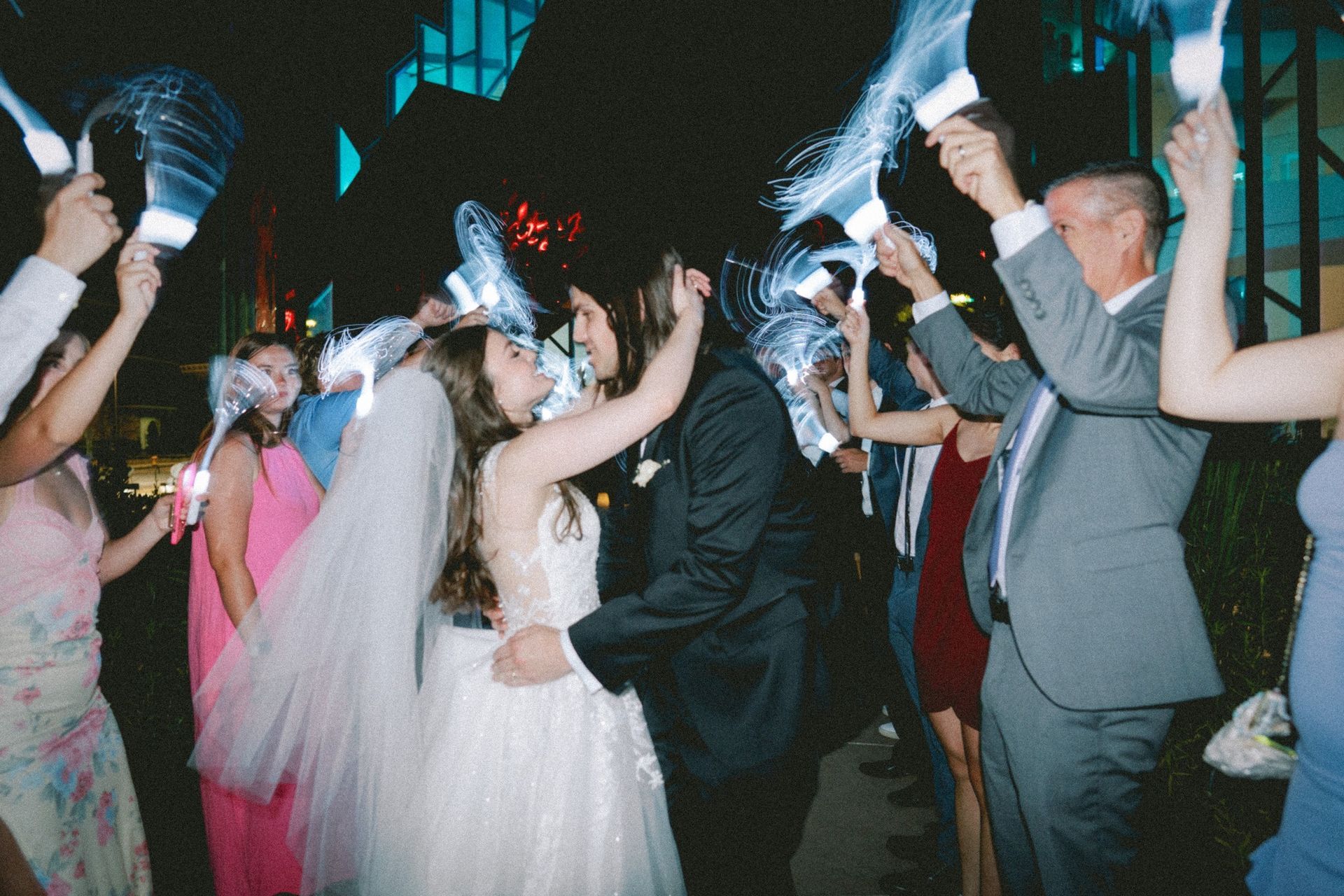 Wedding couple dancing, surrounded by guests holding glowing wands. Nighttime outside a building with colorful lights.