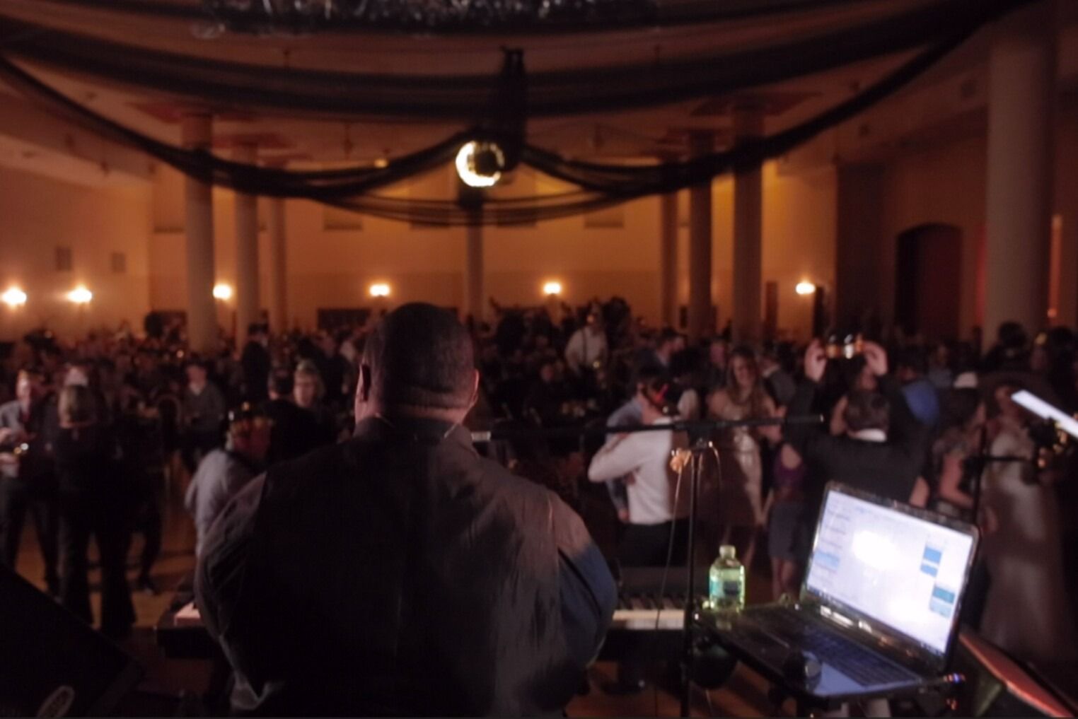 DJ at a crowded event in a large hall with people dancing under dim lighting.