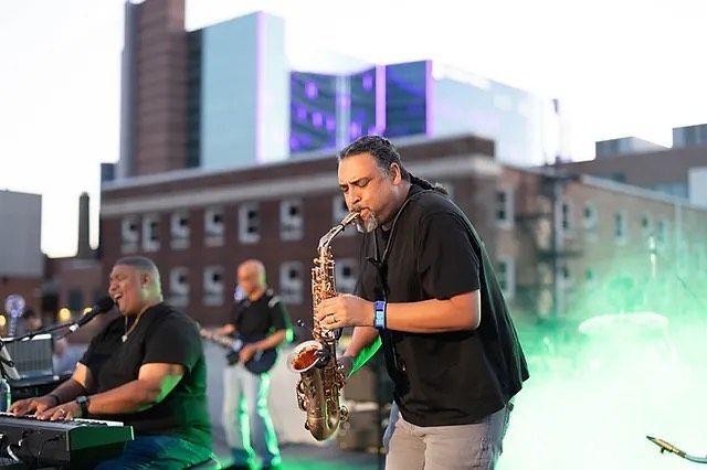 Saxophonist playing on stage with a keyboardist and bassist; buildings in background.