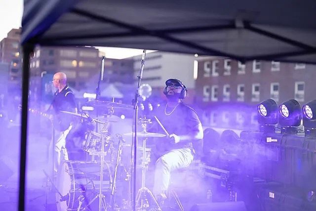 Drummer playing on stage with purple stage lights. Another musician in the background. Outdoor setting.