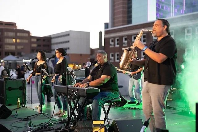 Band performing outdoors: singer, keyboardist, bassist, saxophone player, drummer, and stage.