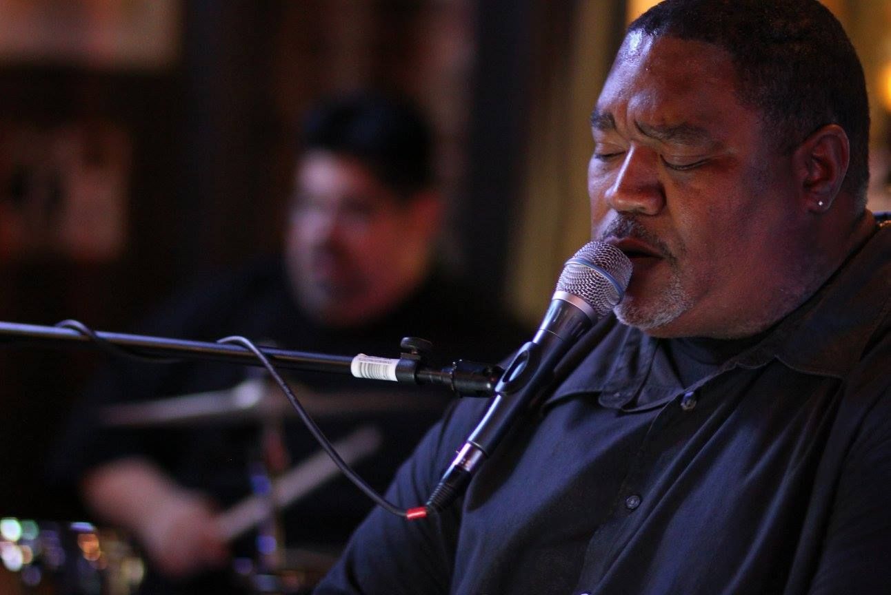 Man singing into microphone, drummer blurred in the background. Low-lit setting.