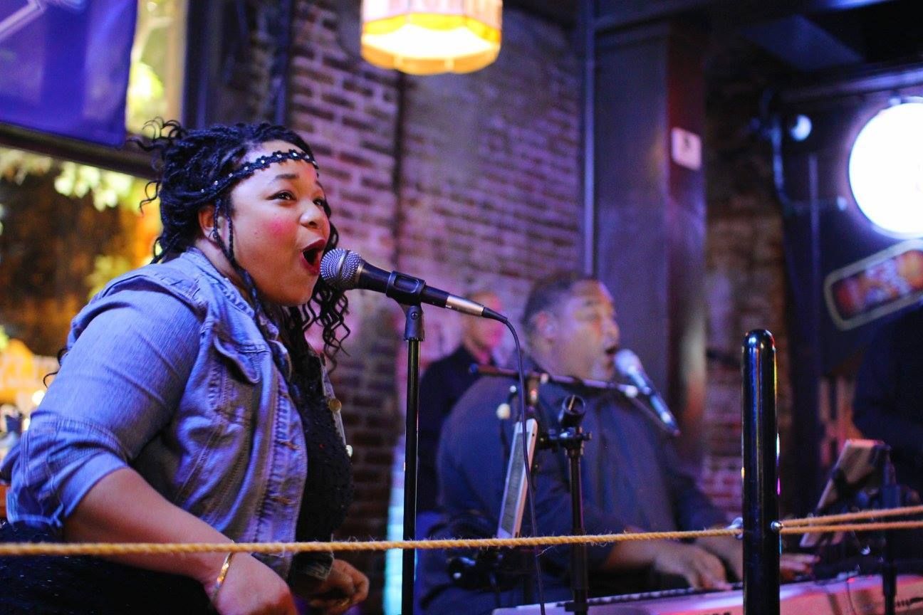 Woman singing into a microphone onstage; man playing keyboard. Dimly lit bar setting.