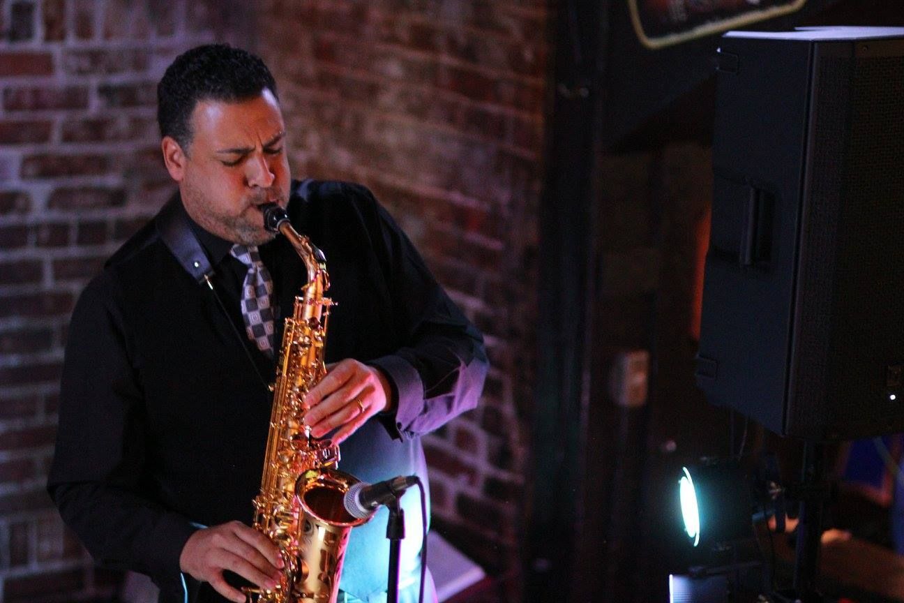 Man playing saxophone in front of a brick wall, wearing a black shirt and tie.