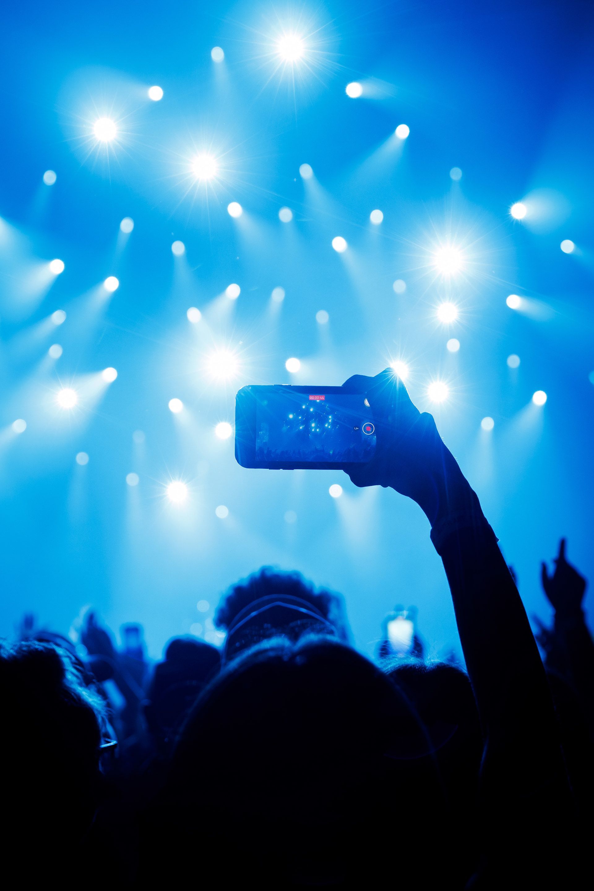 Concert crowd with hands raised holding up phones, lit by bright blue stage lights.