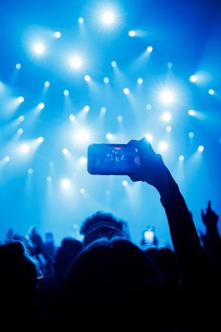 Concert crowd with hands raised holding up phones, lit by bright blue stage lights.